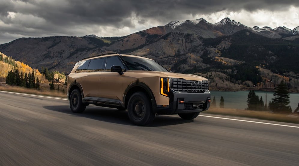 A brown 2027 Kia Telluride SUV driving down a road with mountains and forest in the background