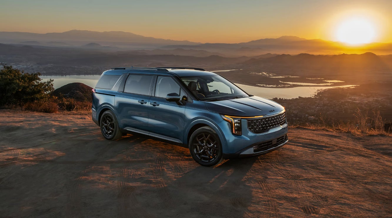 A blue 2026 Kia Carnival MPV parked on a cliff at sunset