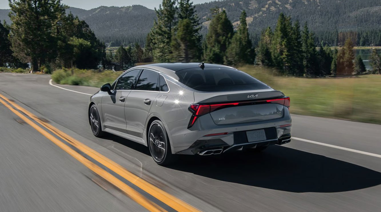 A gray 2026 Kia K5 sedan driving down a road with mountains in the background