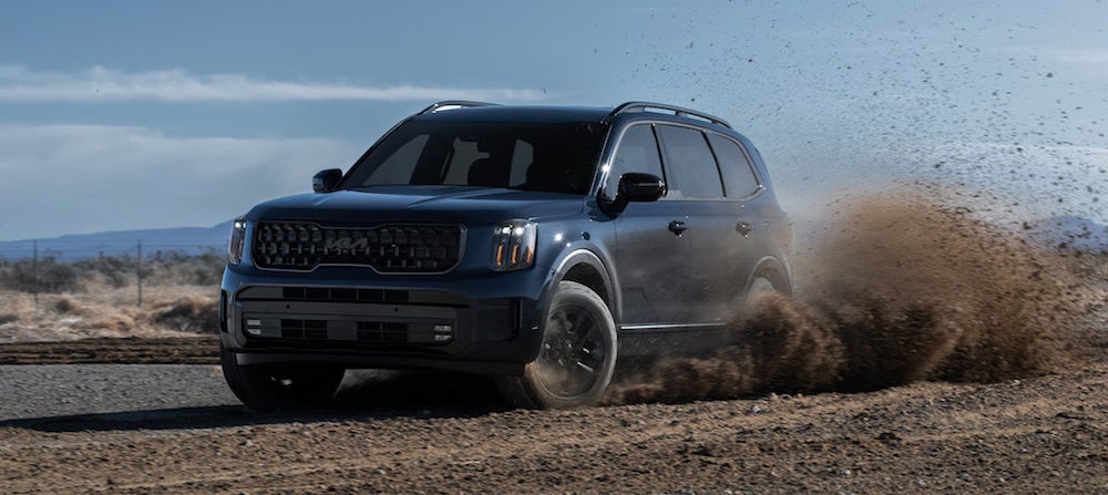 A dark-colored 2025 Kia Telluride SUV driving along a dirt road, kicking up dust 