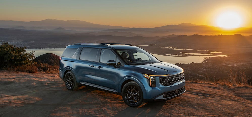 A blue 2026 Kia Carnival MPV parked on a ledge overlooking a river valley at sunset