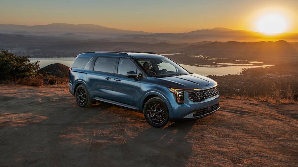 A 2026 Kia Carnival minivan parked on a dirt ledge at sunset, with a body of water and mountains in the background
