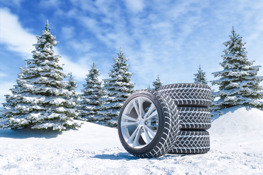 A stack of four snow tires in front of a snowy field with pine trees in the background